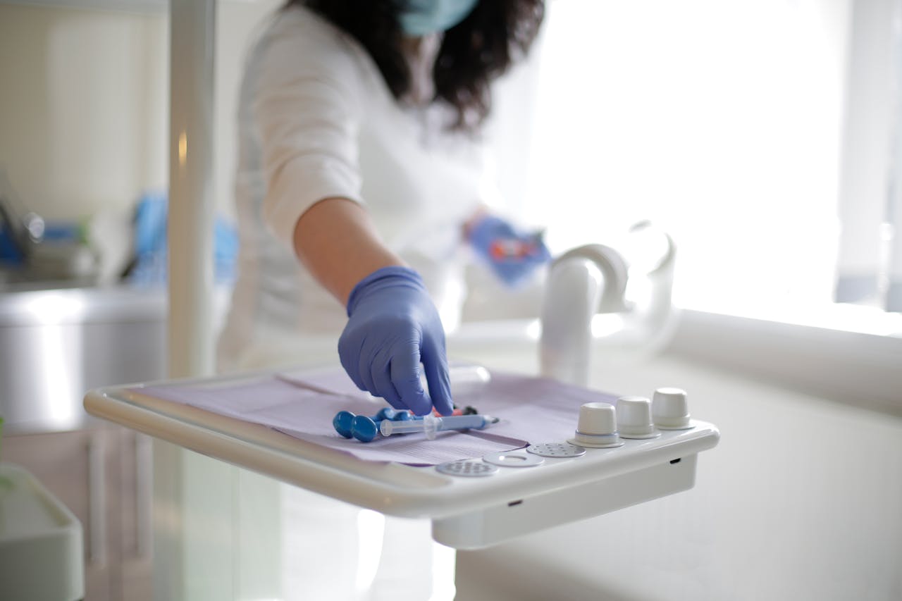 Crop unrecognizable female doctor in latex gloves putting syringe and metal pieces of medical tools on dentist table while standing near window in clinic in sunlight