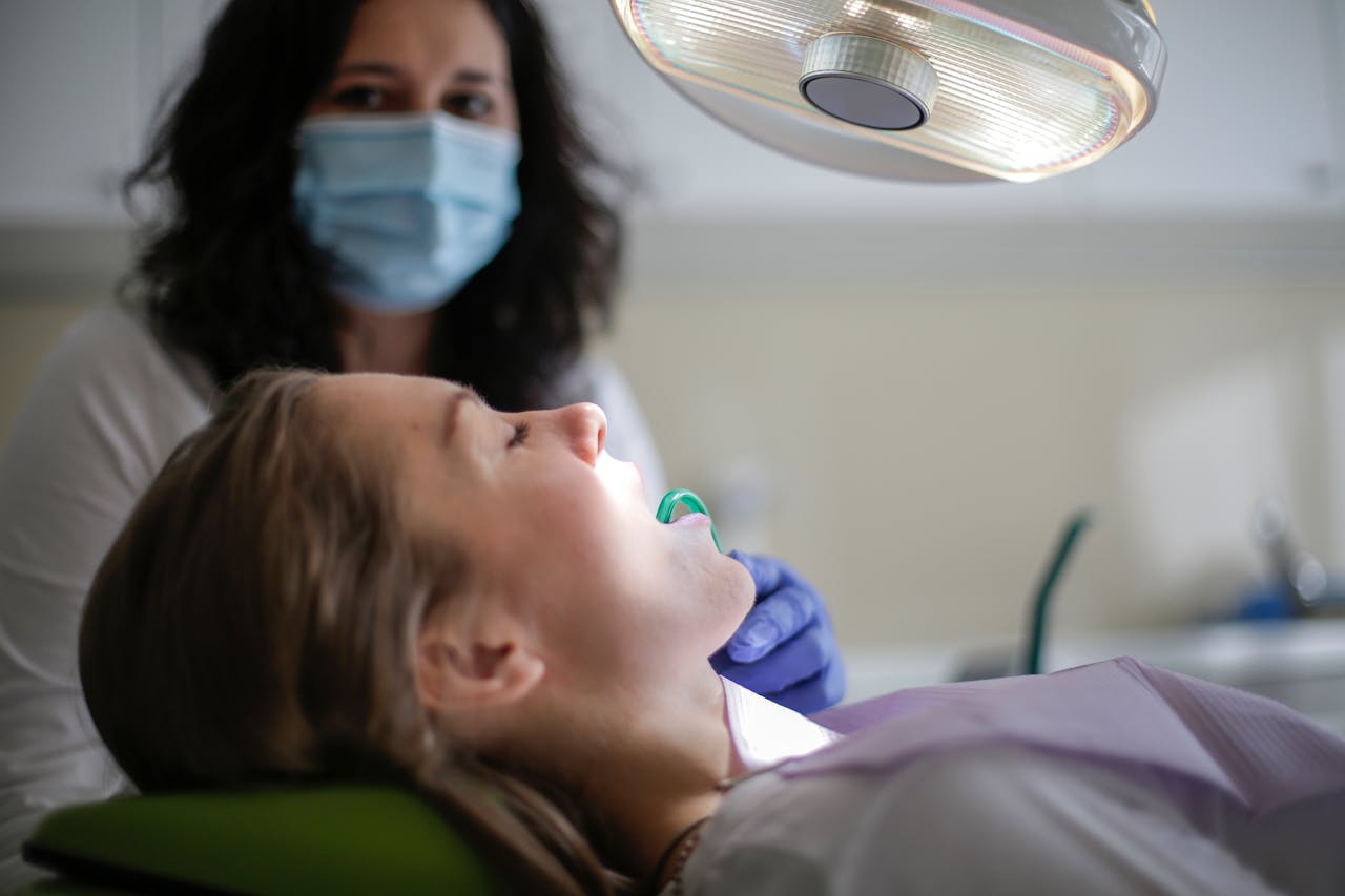 Female patient undergoing dental procedure in clinic. Dentist wearing protective mask conducts examination.
